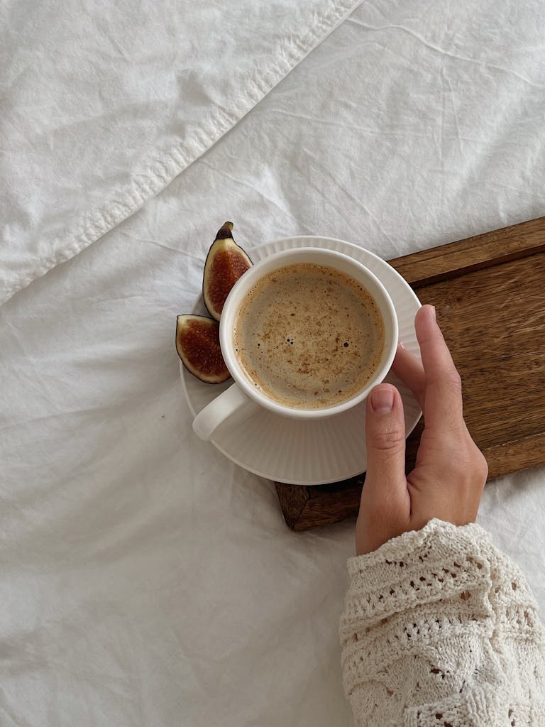 A close-up of a hand holding a coffee cup with figs on a bed, exuding warmth and comfort.