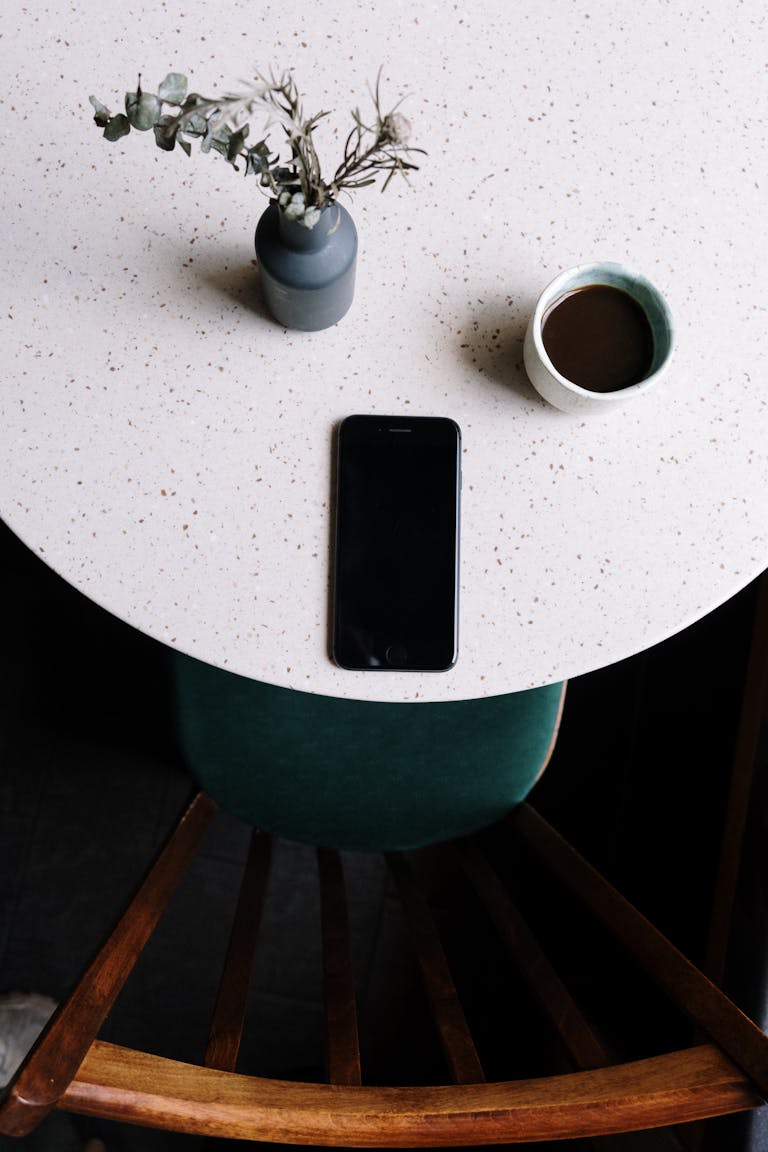 Overhead view of a minimalist setup with a smartphone, coffee cup, and vase on a table.